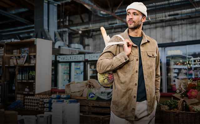 Mann in brauner Jacke und Mütze steht in einem Lagerhaus, trägt einen Brotsack über der Schulter. Im Hintergrund sind Kartons und Regale zu sehen.