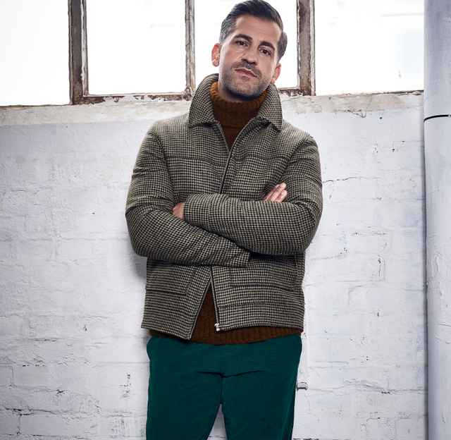 Mann steht vor einer weißen Ziegelwand, gekleidet in eine karierte Jacke, braunen Pullover und grüne Hose, mit verschränkten Armen.