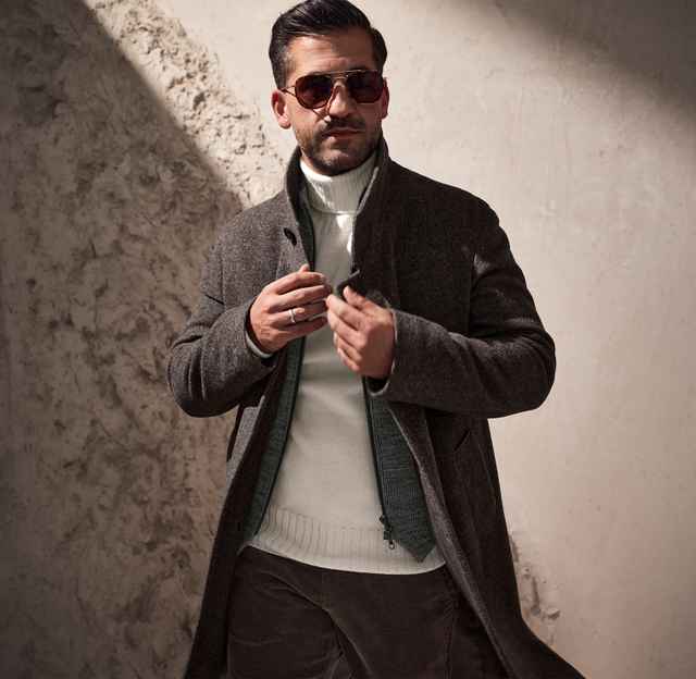 Mann mit Sonnenbrille trägt einen winterlichen, grauen Mantel über einem weißen Rollkragenpullover, steht vor einer schattigen Wand.