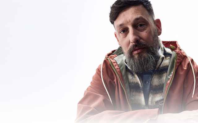 Mann mit Bart in roter Jacke, vor hellem Hintergrund sitzend.