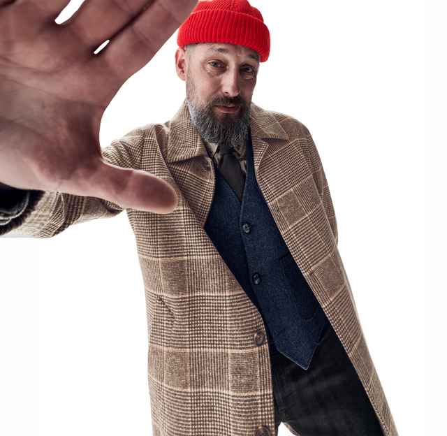 Mann mit roter Mütze und kariertem Mantel schaut in die Kamera und hebt seine Hand zum Stoppzeichen. Hintergrund ist weiß.