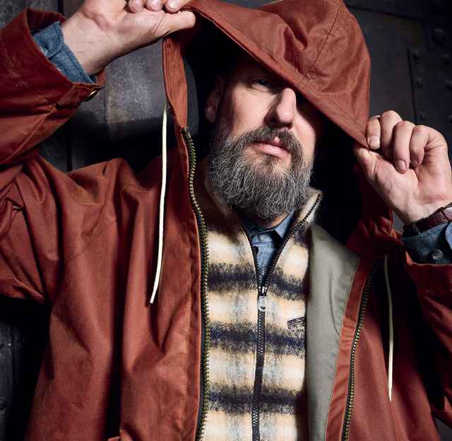 Mann mit grauem Bart trägt eine braune Jacke mit Kapuze und eine karierte Weste, hebt die Kapuze mit den Händen an.