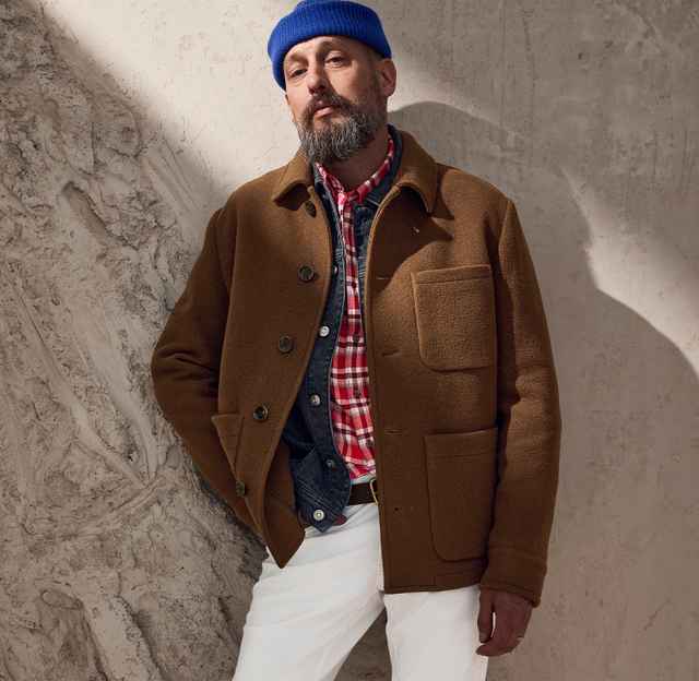 Mann mit blauem Mütze, brauner Jacke, kariertem Hemd und weißer Hose lehnt an einer strukturierten Wand, in einem modischen, lässigen Stil.