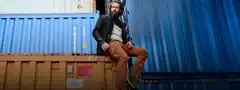 Mann mit Bart sitzt lässig auf einem rostigen Container vor blauen weiteren Containern im Hintergrund. Er trägt braune Hosen und dunkle Jacke.