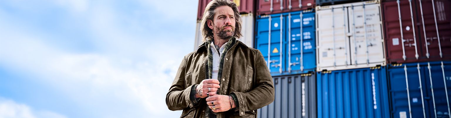 Ein Mann mit brauner Jacke steht vor aufgestapelten Containern unter einem blauen Himmel.