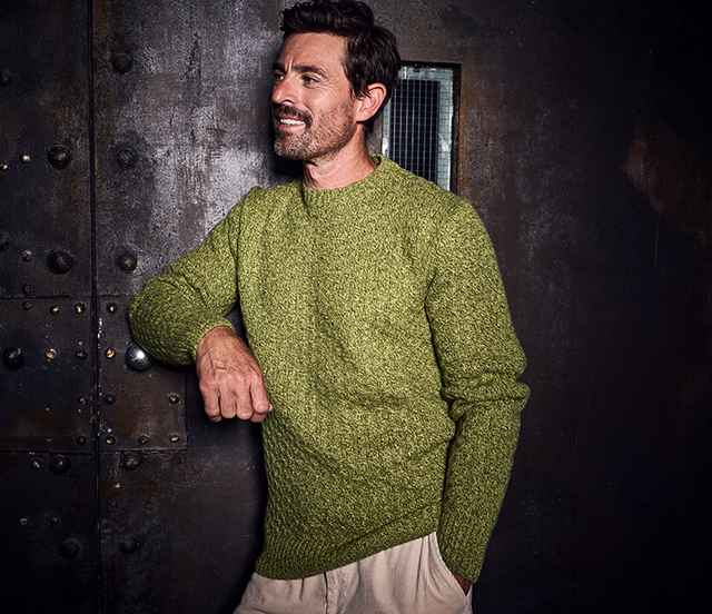 Mann mit grünem Pullover lehnt lächelnd an einer dunklen Metalltür.
