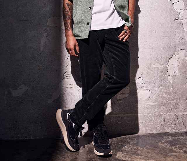 Person lehnt an einer Wand, trägt schwarze Hose, weißes T-Shirt und Skelettmantel mit dunklen Sneakers. Karge Umgebung mit Betonboden.