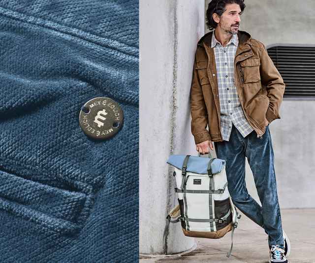 Mann in brauner Jacke und karierter Hemd lehnt an Wand, trägt große Rucksack. Detailaufnahme eines Knopfs auf blauer Stoffoberfläche links.