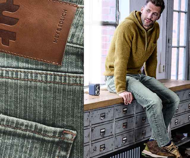 Ein Mann in gelbem Hoodie sitzt auf einem Schrank neben einer Nahaufnahme einer Jeans mit Lederetikett.