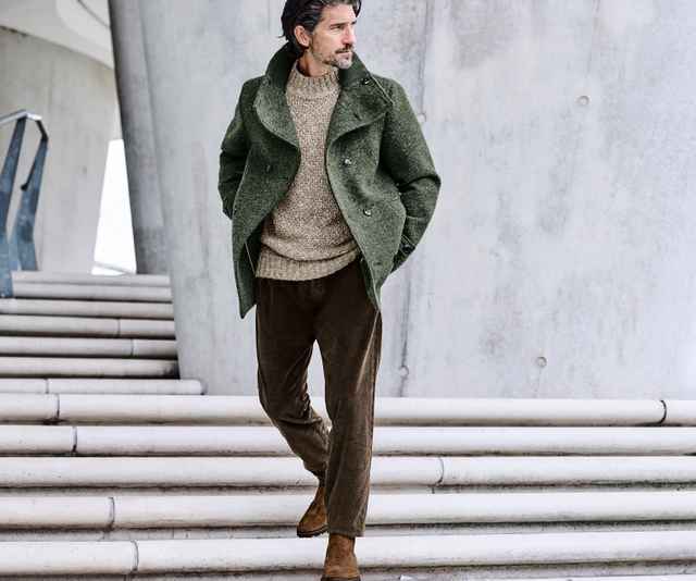 Mann mit grünem Mantel und beige Pullover geht eine Treppe hinunter. Hintergrund: moderne Betonstruktur.