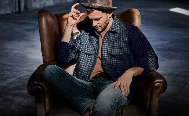 Mann sitzt in Ledersessel, trägt blau kariertes Hemd, Jeans und Hut, auf grauem Hintergrund.