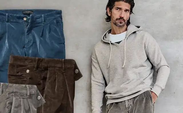 Mann in grauem Hoodie steht vor einer Wand mit drei gefalteten Hosen in Blau, Braun und Grau.