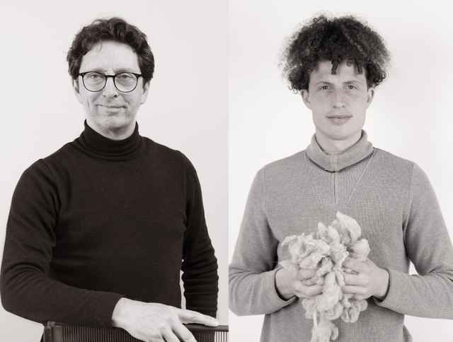 Zwei Männer stehen nebeneinander, einer in schwarzem Rollkragenpullover, der andere in grauem Pullover, hält Wolle in den Händen.